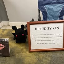 A plush black toy with red cheeks and ears, a framed sign with text, and a blue and gray backpack on a wooden surface.