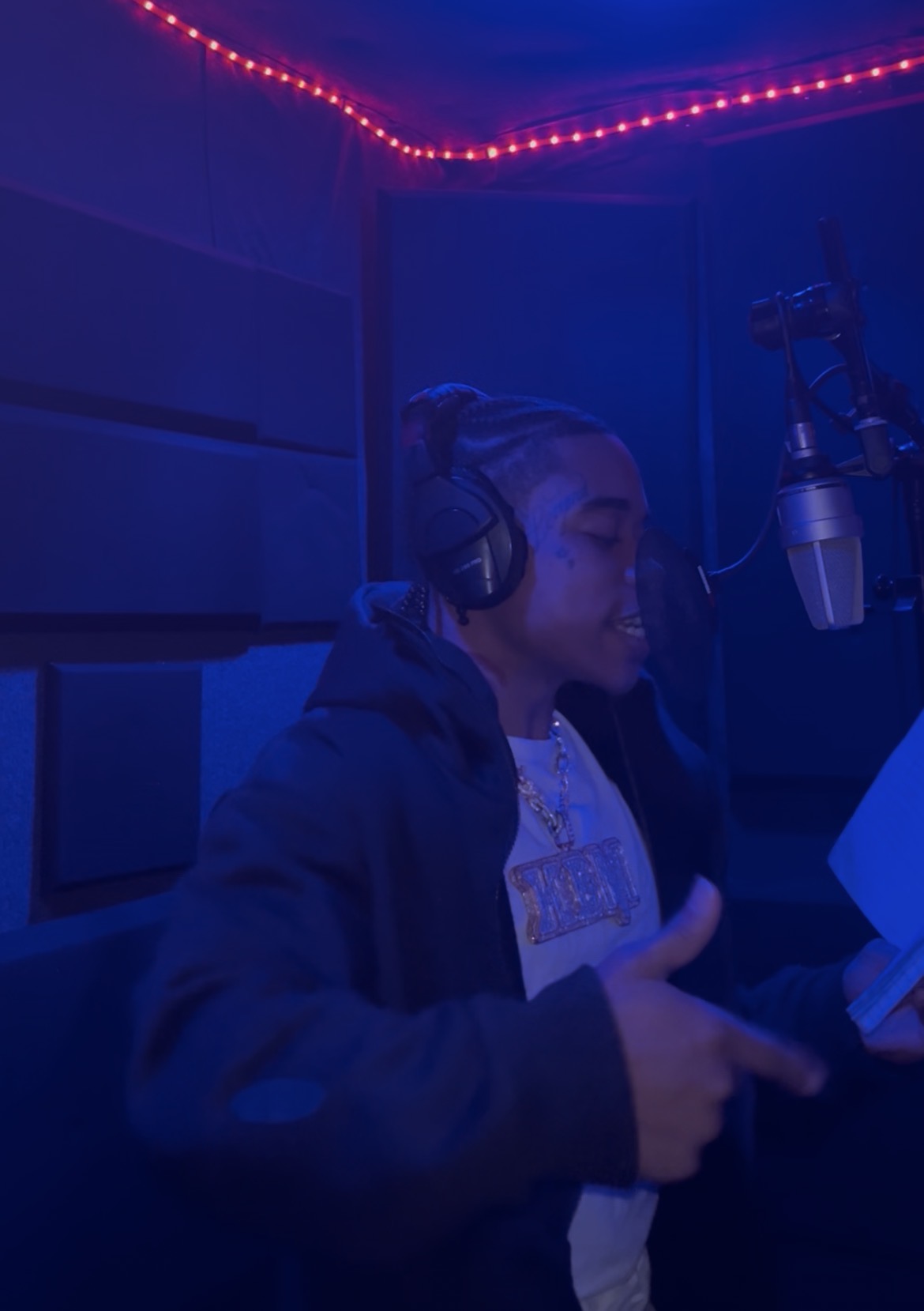 Person in a recording studio with headphones speaking into a microphone, illuminated by blue and red lights.
