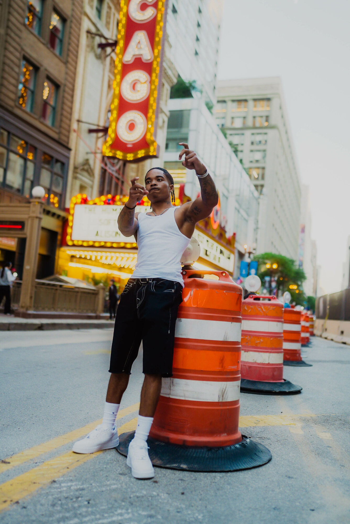 Young man standing on city street with orange traffic barrels, tall buildings, and a neon Chicago sign in the background.
