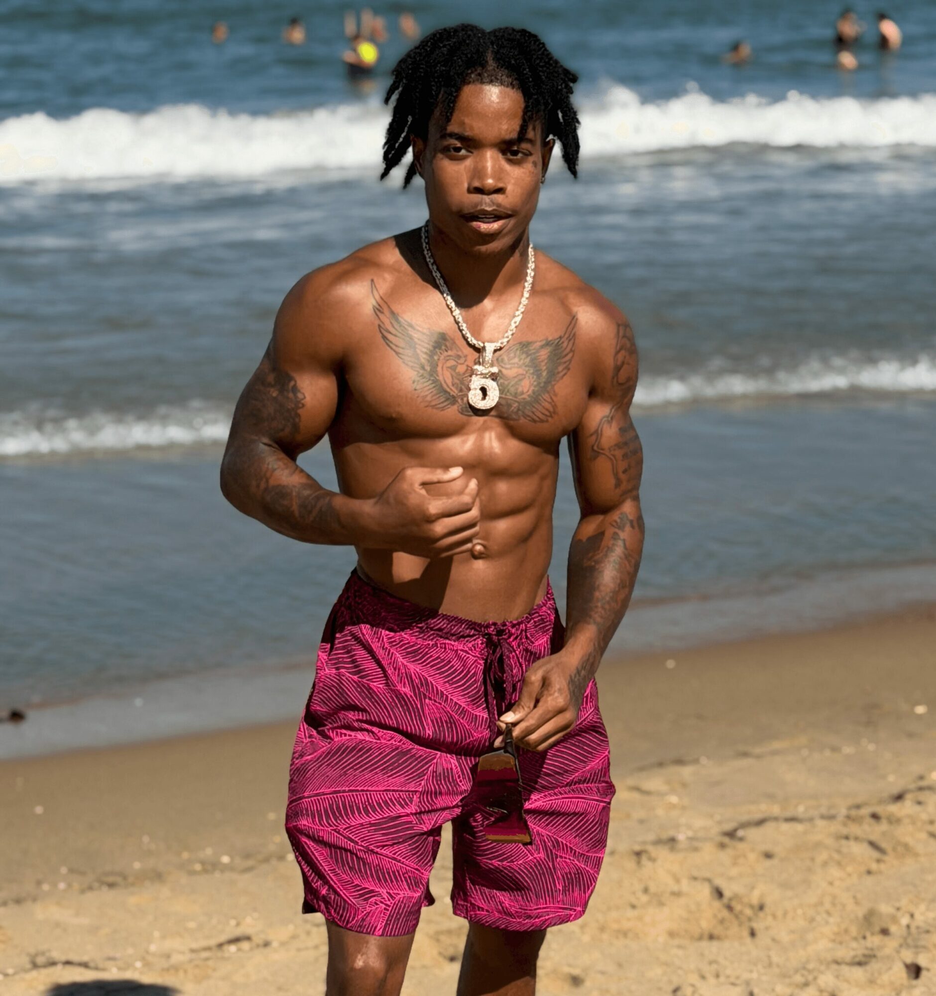 Young man with tattoos and dreadlocks standing on a beach, wearing pink shorts, with ocean and people in the background.