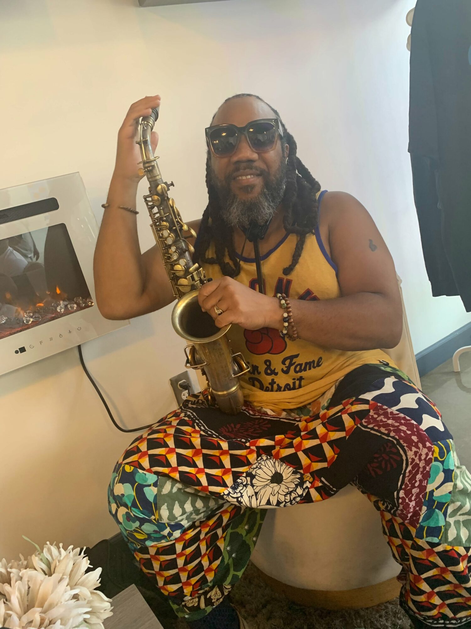 Person with long hair, sunglasses, and a beard sitting on a toilet holding a saxophone, wearing colorful patterned pants and a yellow top.