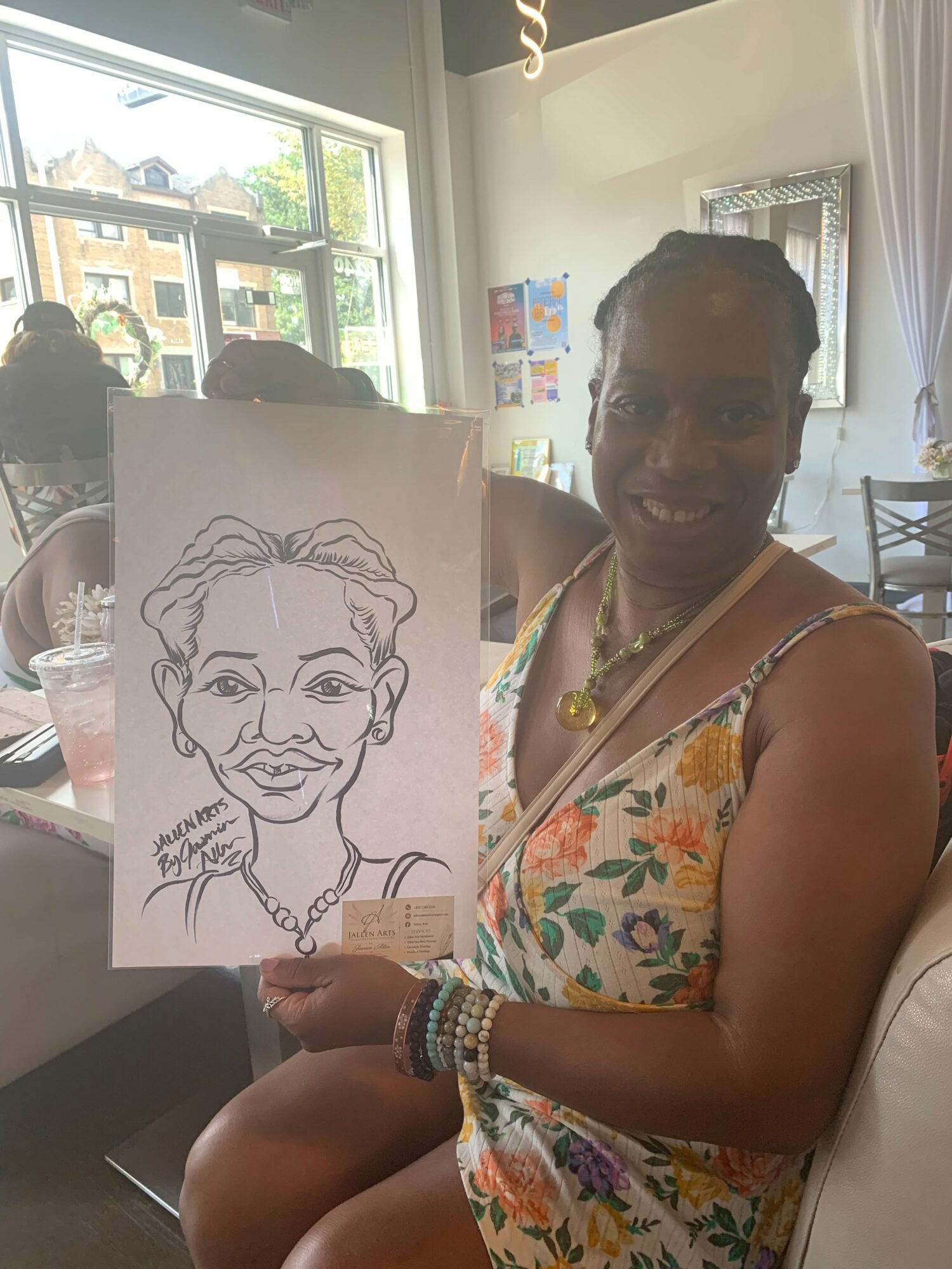 Woman in floral dress holding a caricature drawing of herself, smiling, in a bright room with large windows.