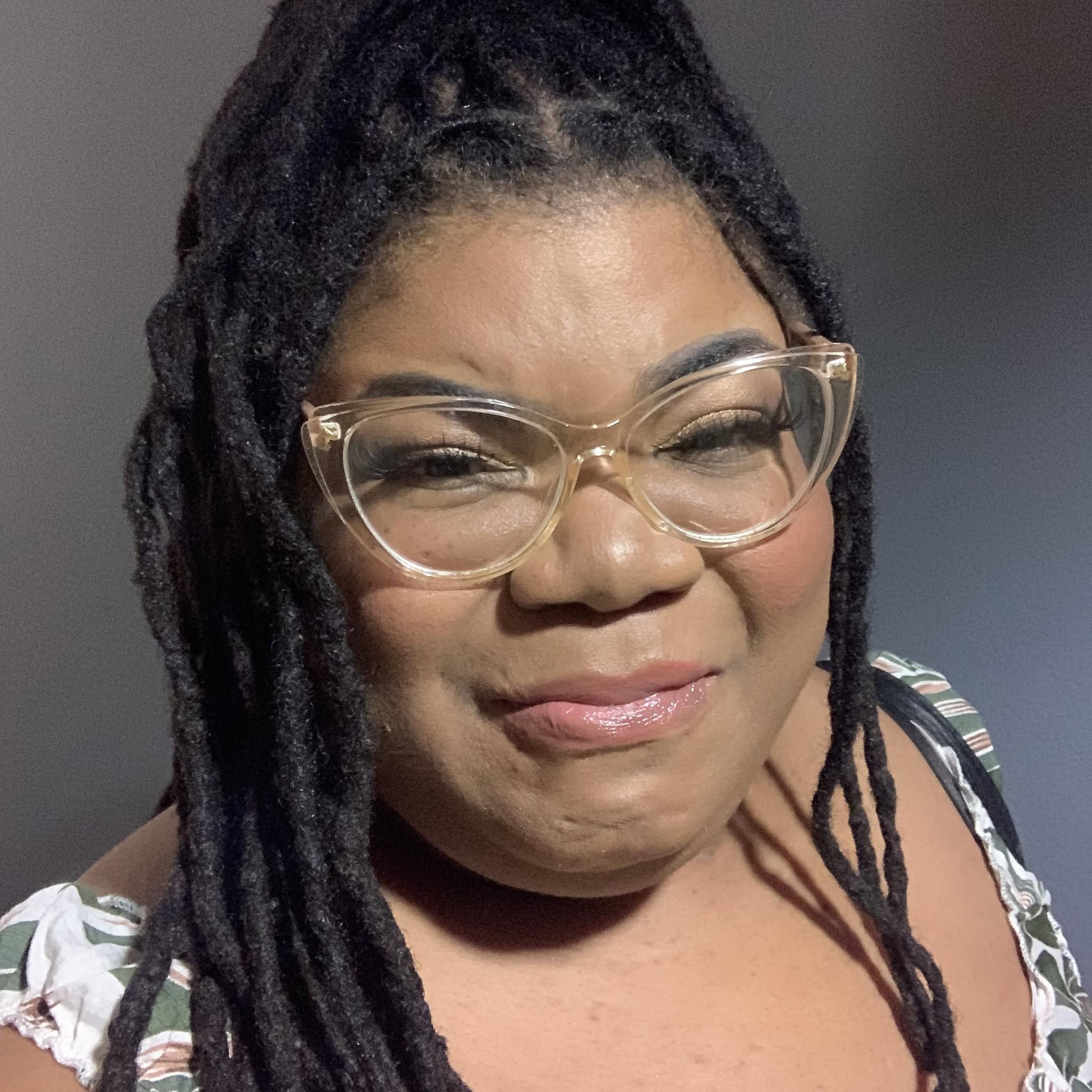 Woman with glasses and dreadlocks smiling, wearing a patterned top, indoors with a neutral background.