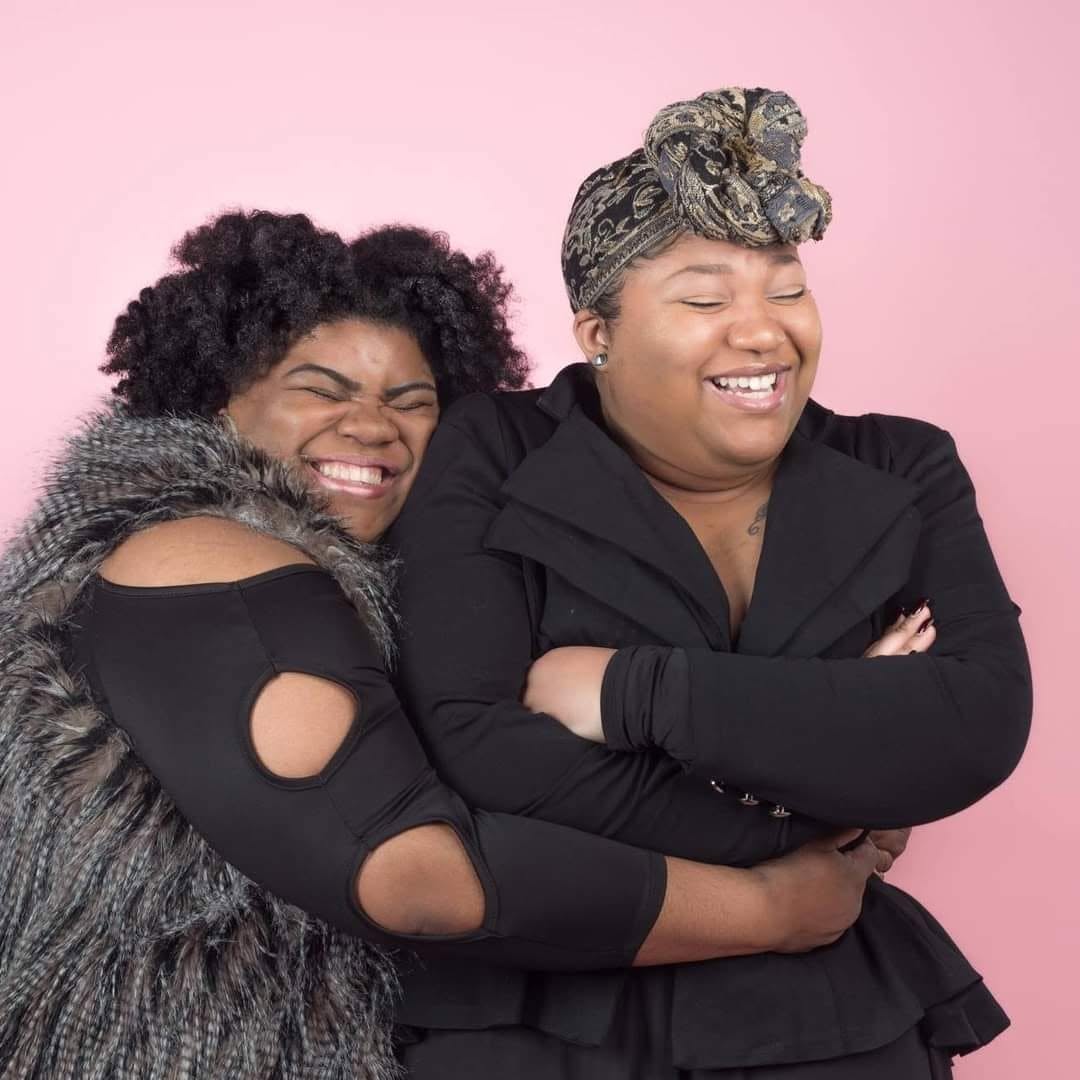 Two women smiling and hugging against a pink background, one with curly hair and the other with a headscarf, arms crossed.