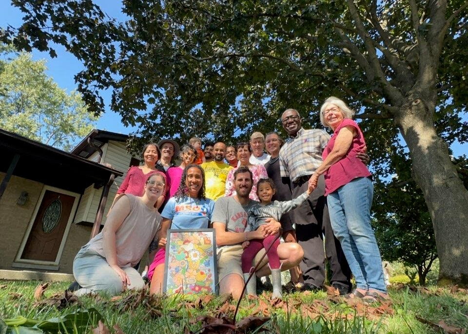 Group of people outdoors under a large tree, smiling and holding hands, with a house in the background.