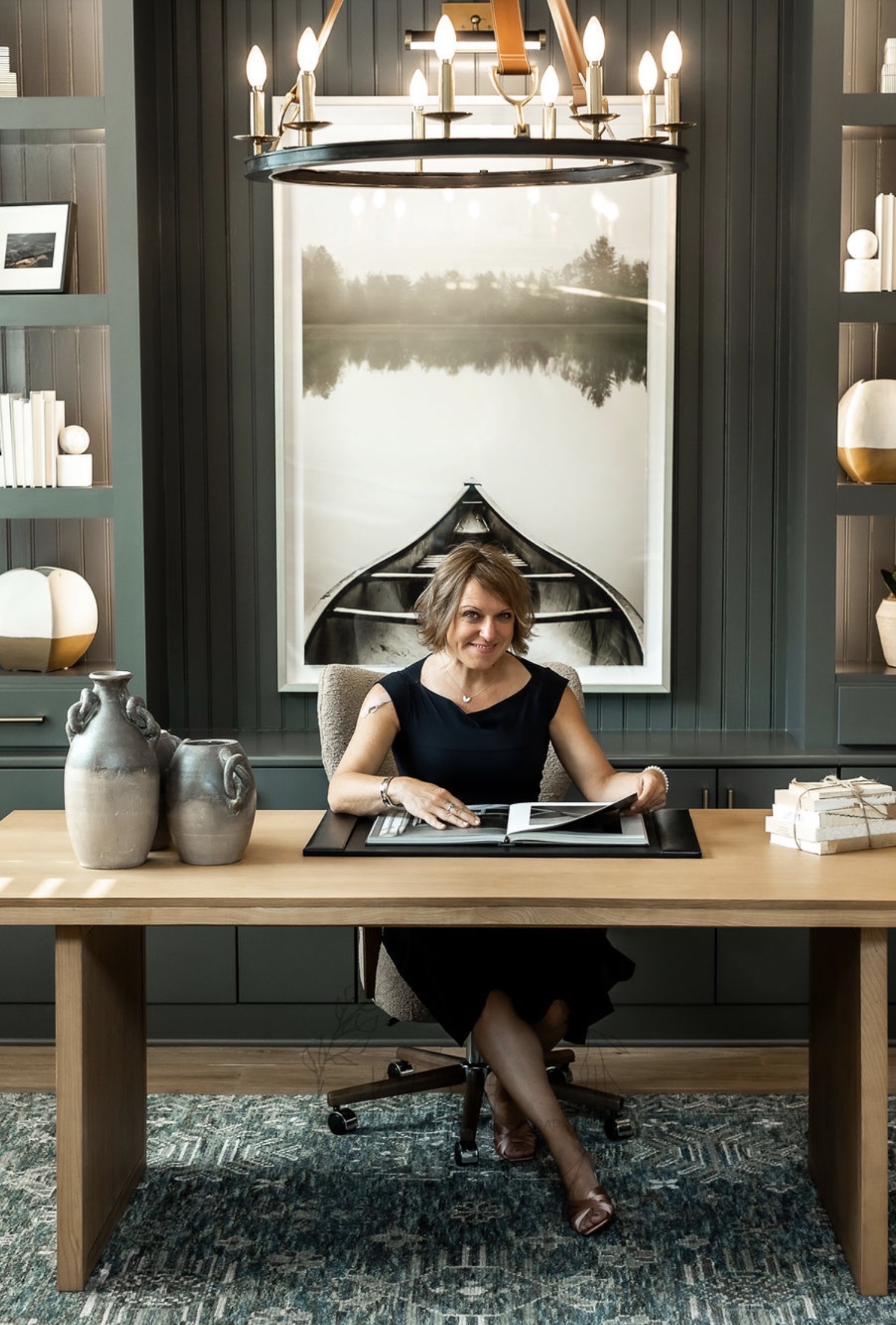 Woman sitting at a desk in a room with a large window, a chandelier, and decorative vases, reading a book.
