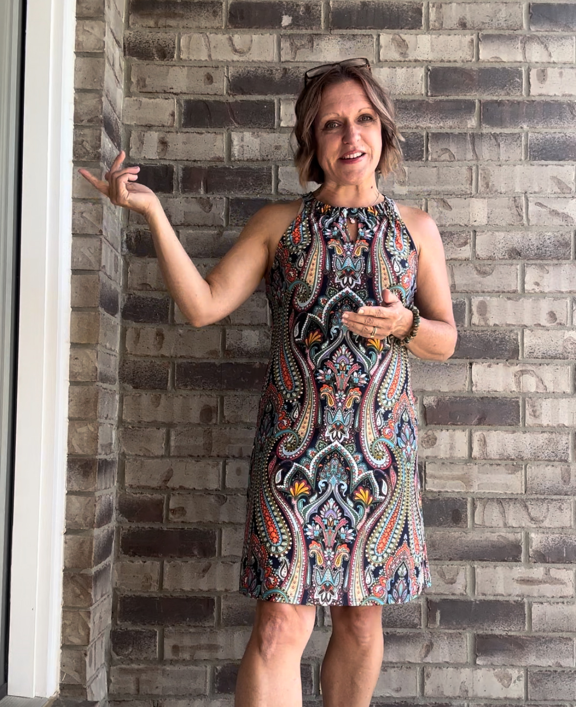 Woman in a colorful patterned dress standing against a brick wall, smiling, with her left arm raised and holding a phone.