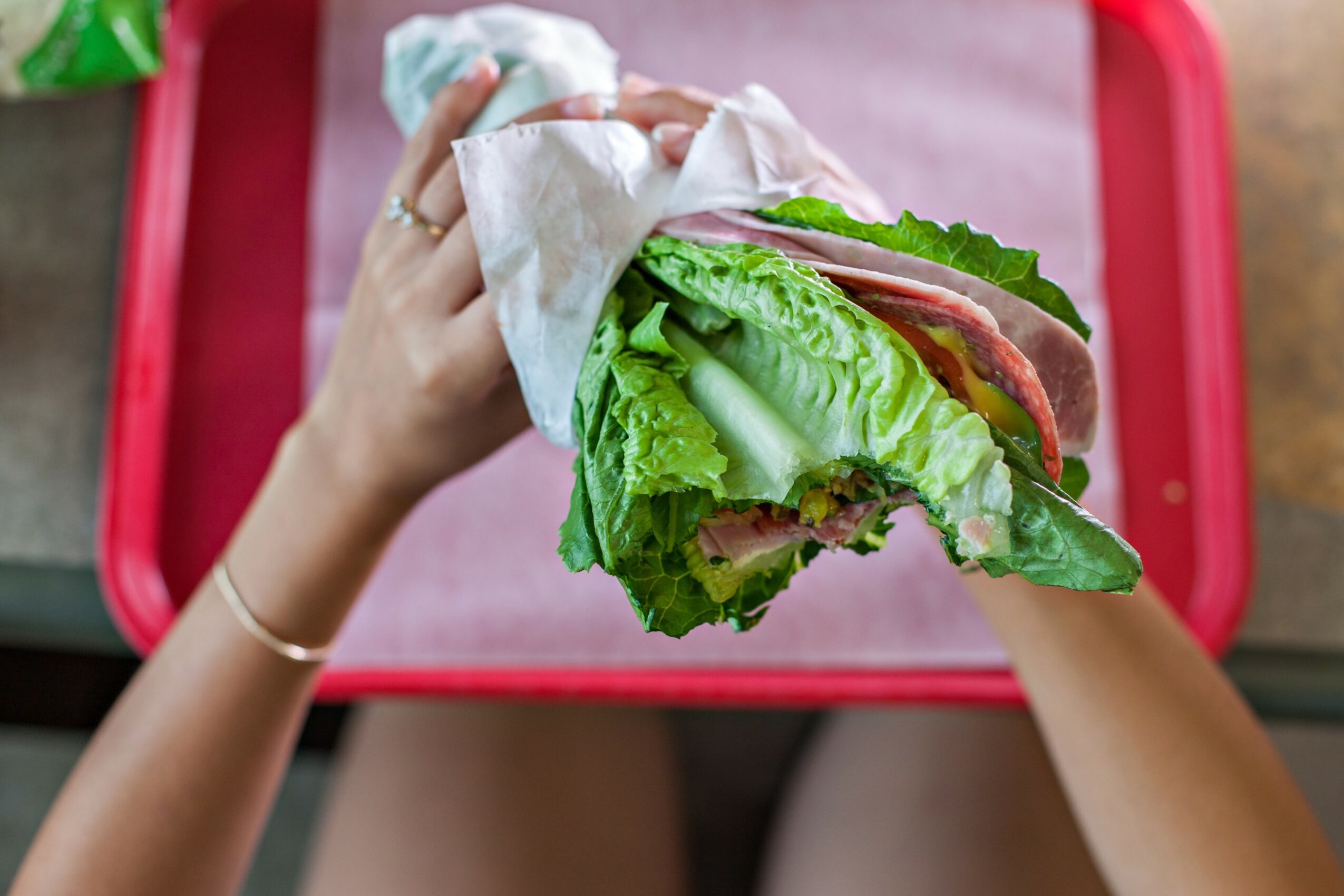 Person holding a large sandwich with lettuce, tomato, and meat, wrapped in paper, over a pink tray.