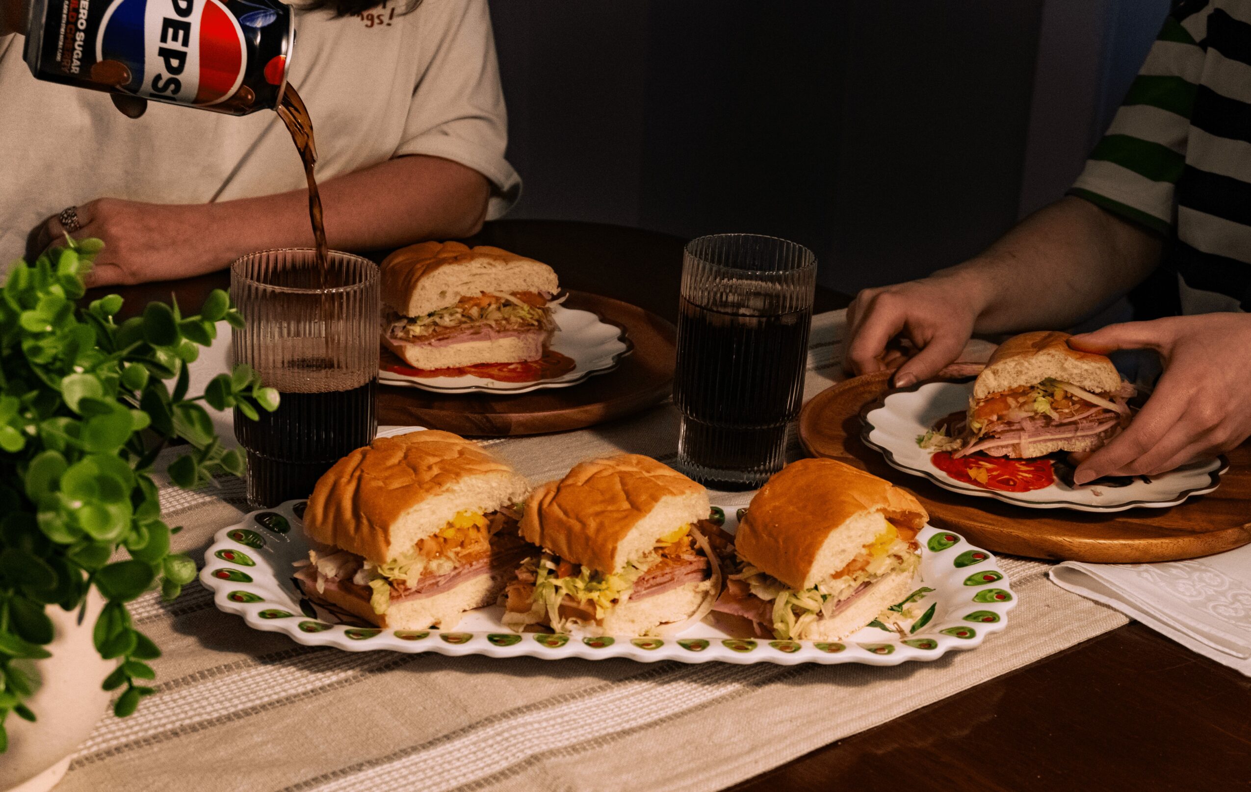 Four sandwiches on a plate, two people with sandwiches and drinks at a table, a plant on the side.