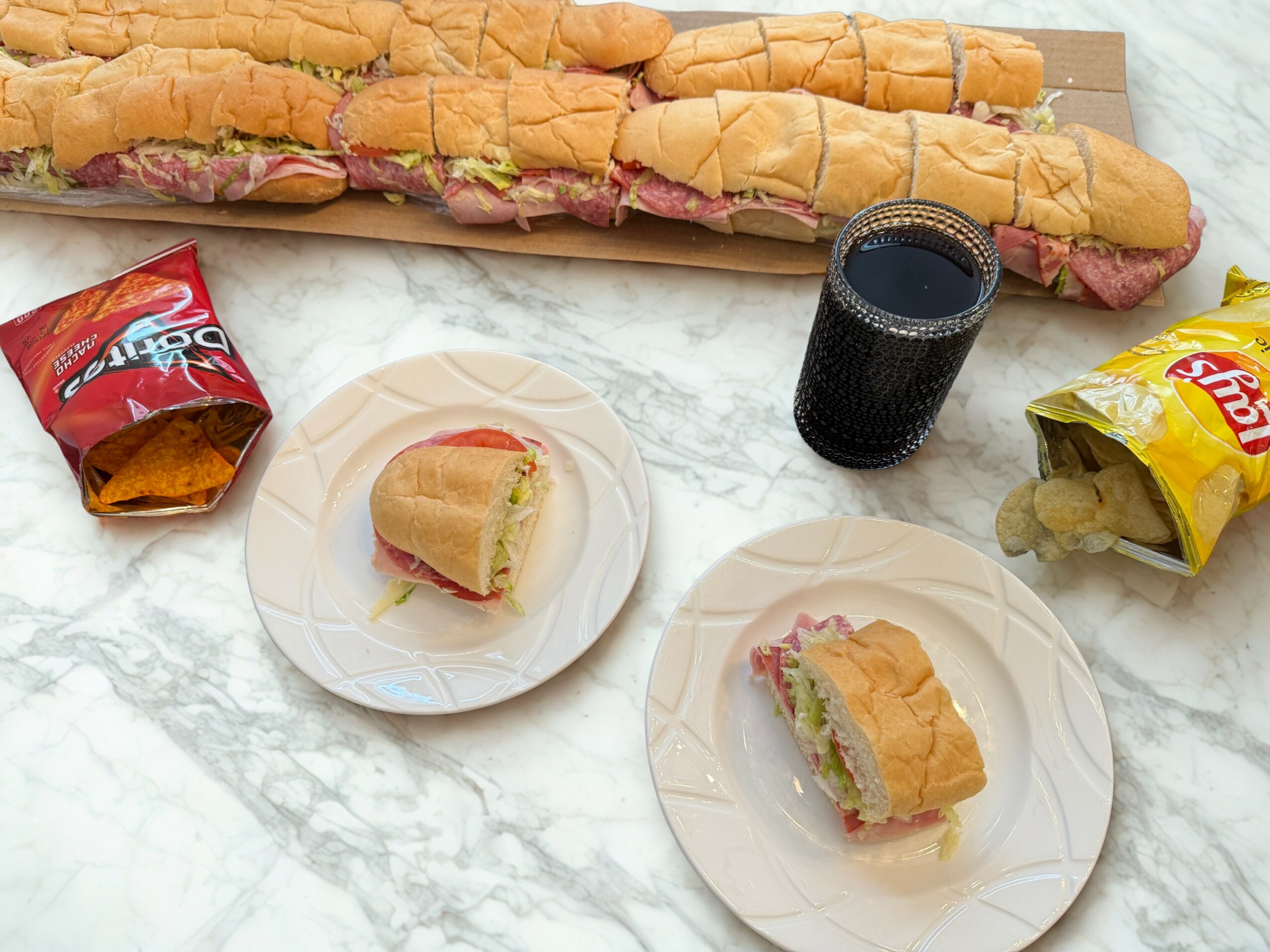 Two sandwiches on plates, a glass of soda, and snack bags on a marble surface.
