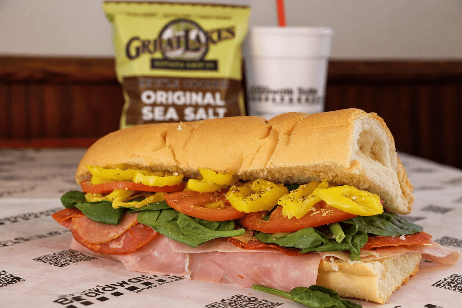 Sub sandwich with ham, lettuce, tomato, pickles, and yellow mustard on a paper wrapper, with snack bag and drink in background.
