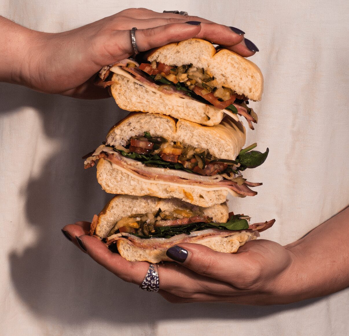 Two hands holding a stack of four sandwiches with various fillings, against a light background.