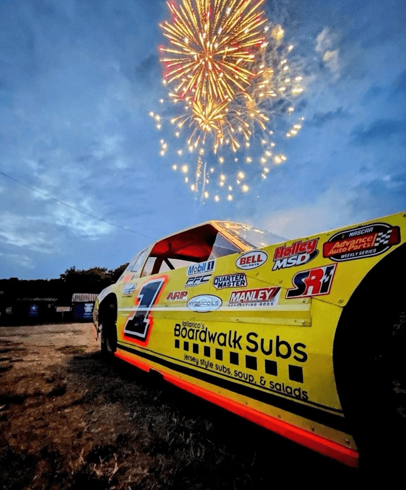 Race car with sponsor logos parked on dirt ground, fireworks in the sky above, blue sky with clouds.