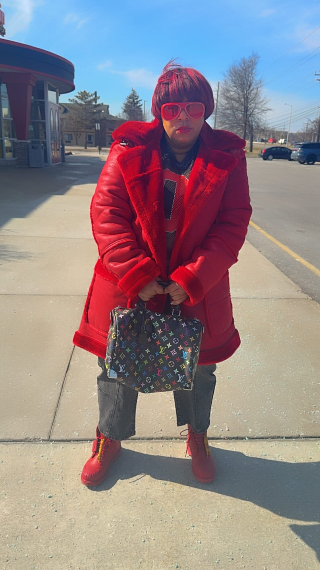 Person standing outdoors on sidewalk wearing red coat, sunglasses, holding a patterned bag, with blue sky and trees in background.