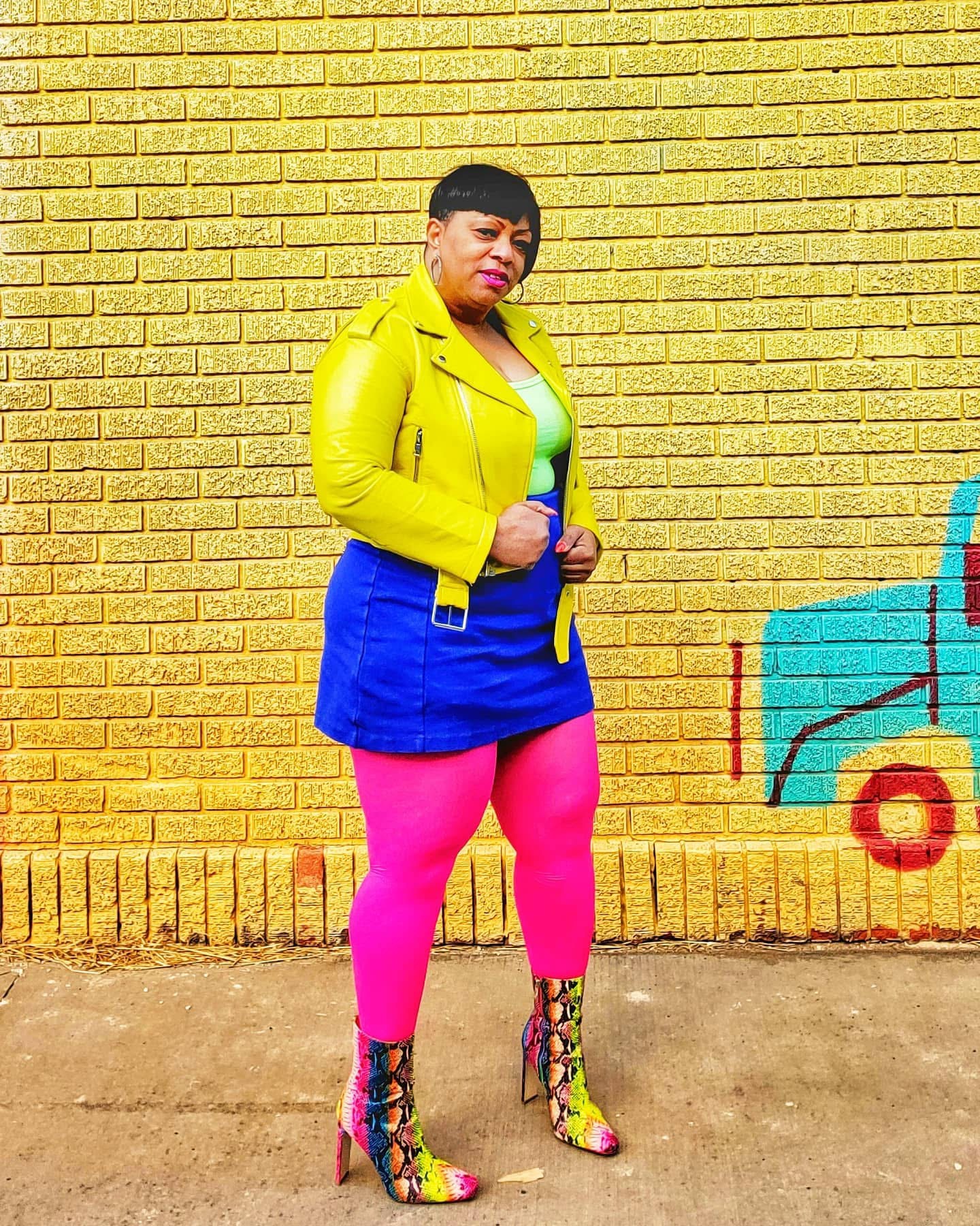 Person standing against a yellow brick wall, wearing a yellow jacket, blue skirt, pink tights, and colorful high heels.