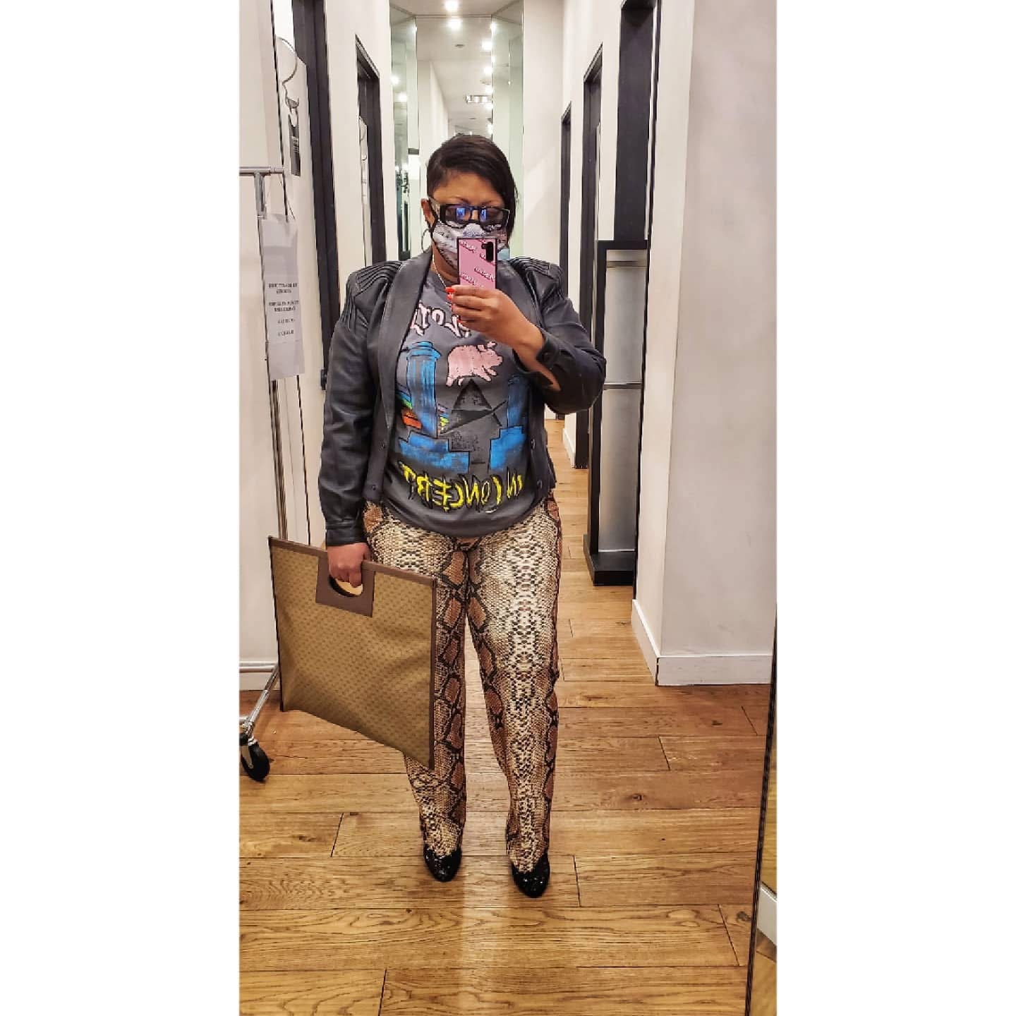 Person taking a mirror selfie in a hallway, wearing glasses, a graphic t-shirt, patterned pants, and carrying a large bag.