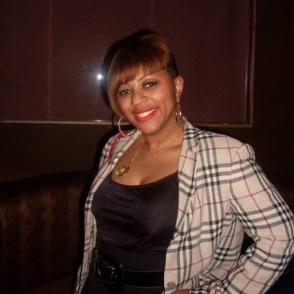 Woman with short hair smiling, wearing a plaid blazer over a black top, in a dimly lit room.