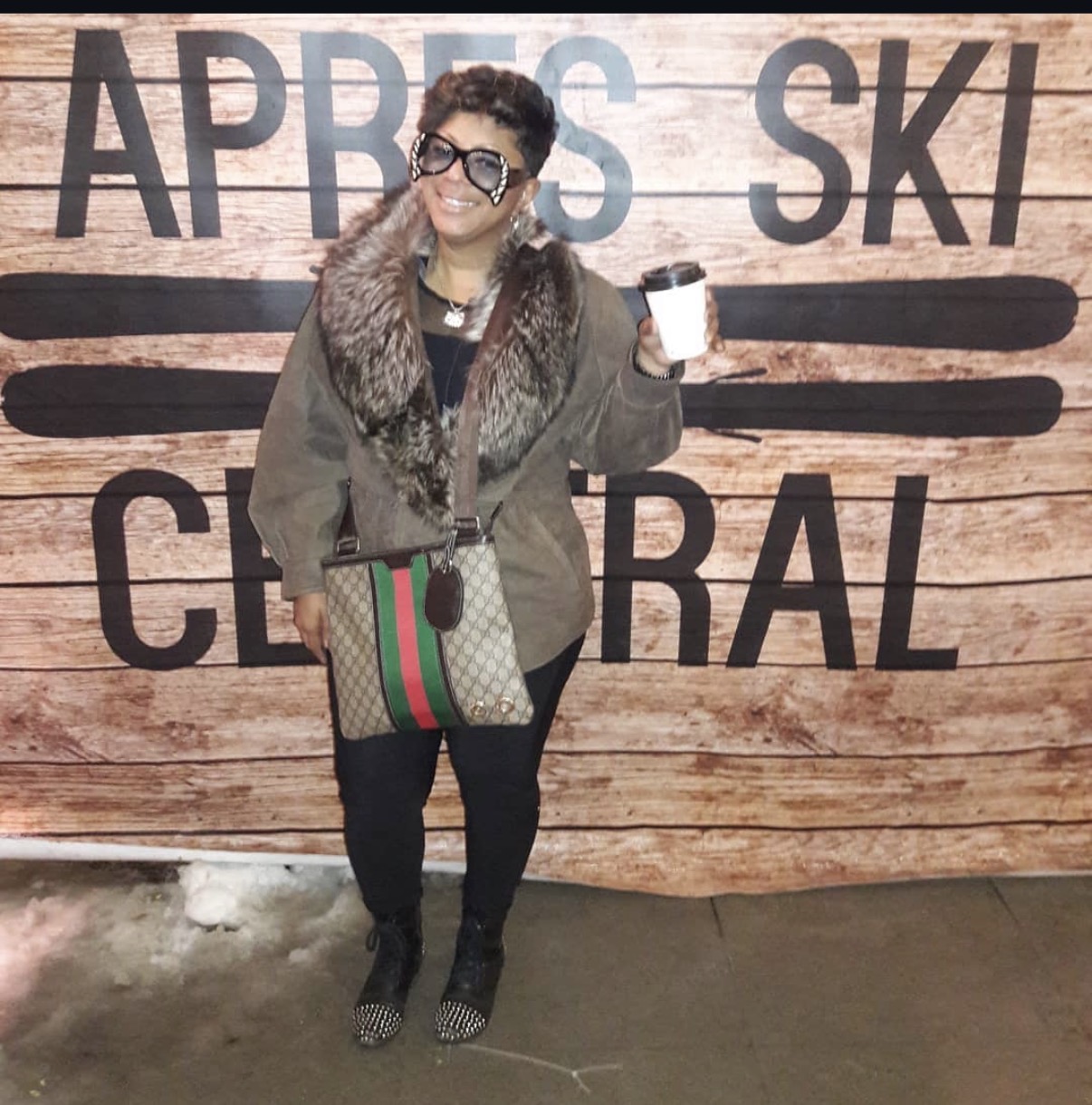 Woman with sunglasses and fur collar coat holding a cup, standing in front of a wooden sign with large text.