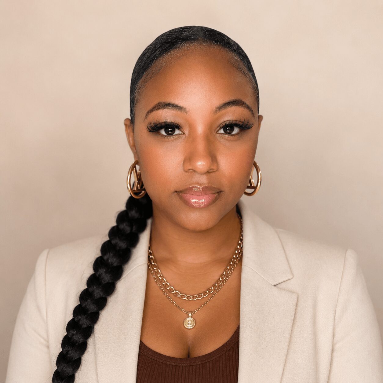 Portrait of a woman with braided hair, gold hoop earrings, layered necklaces, wearing a beige blazer and brown top, neutral background.