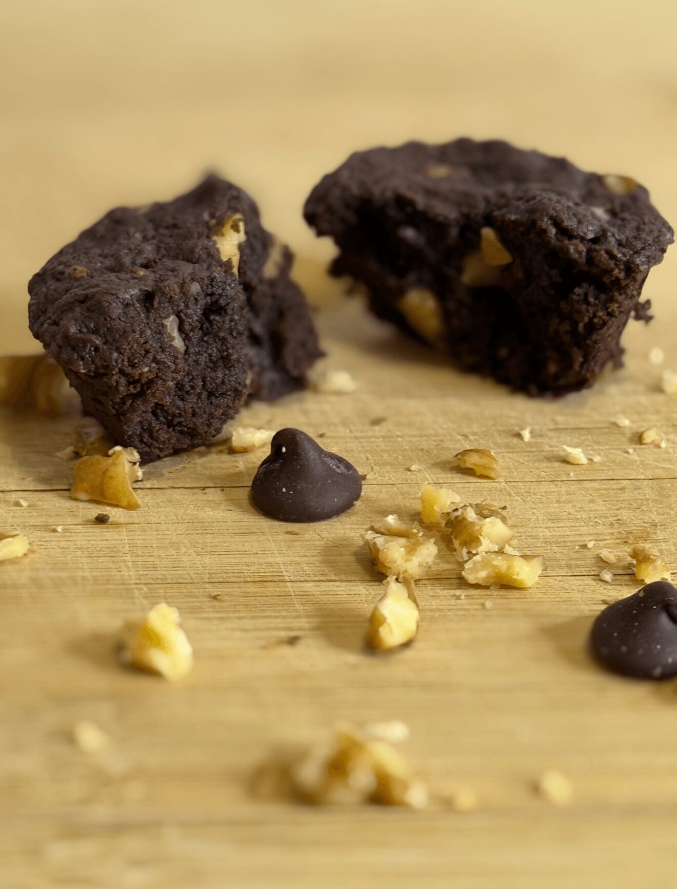 Two chocolate brownies with nuts on a wooden surface, with scattered nuts and chocolate chips around them.