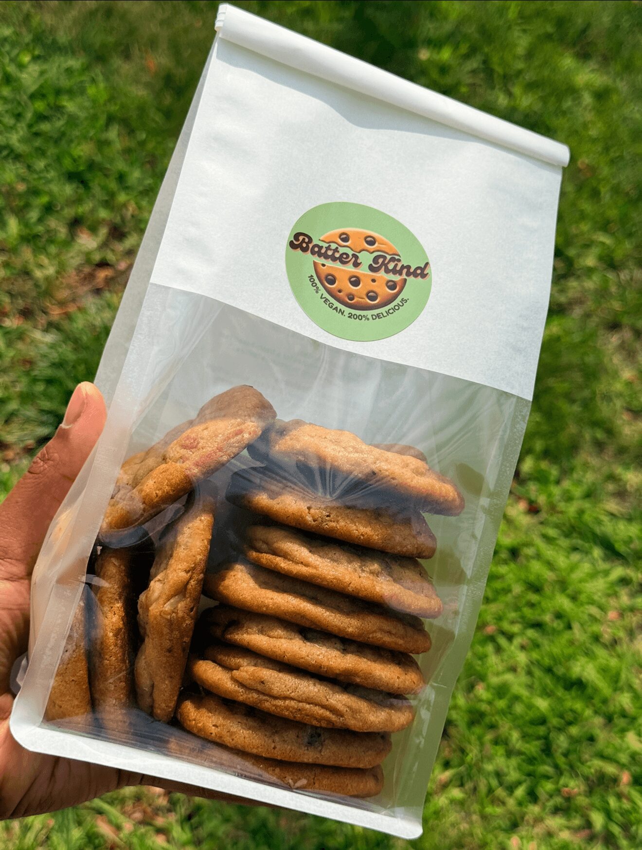 Hand holding a clear bag of cookies with a green and orange logo on white paper top, outdoors with grass background.