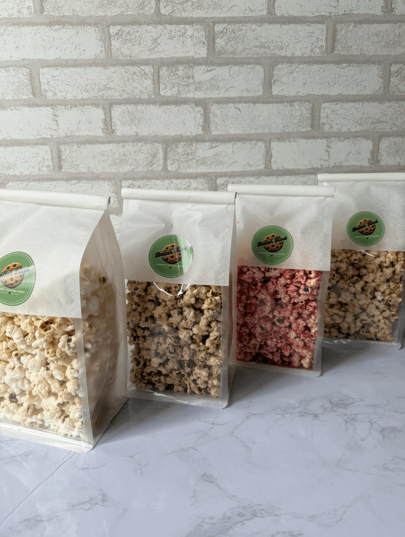 Four clear plastic bags with colorful labels, filled with different types of popcorn, placed on a white surface against a brick wall.