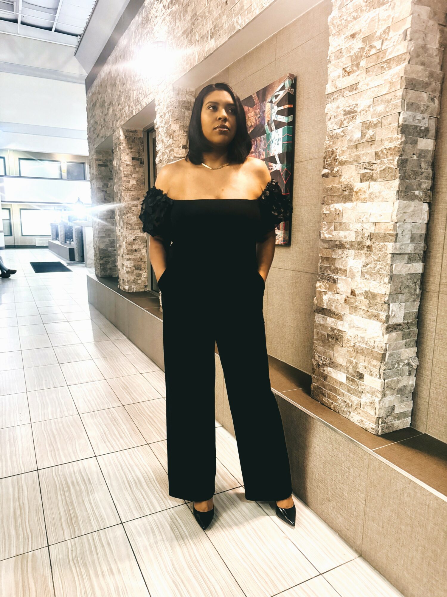 Woman in black off-shoulder jumpsuit standing indoors near brick wall, looking to the side, with sunlight and artwork behind her.