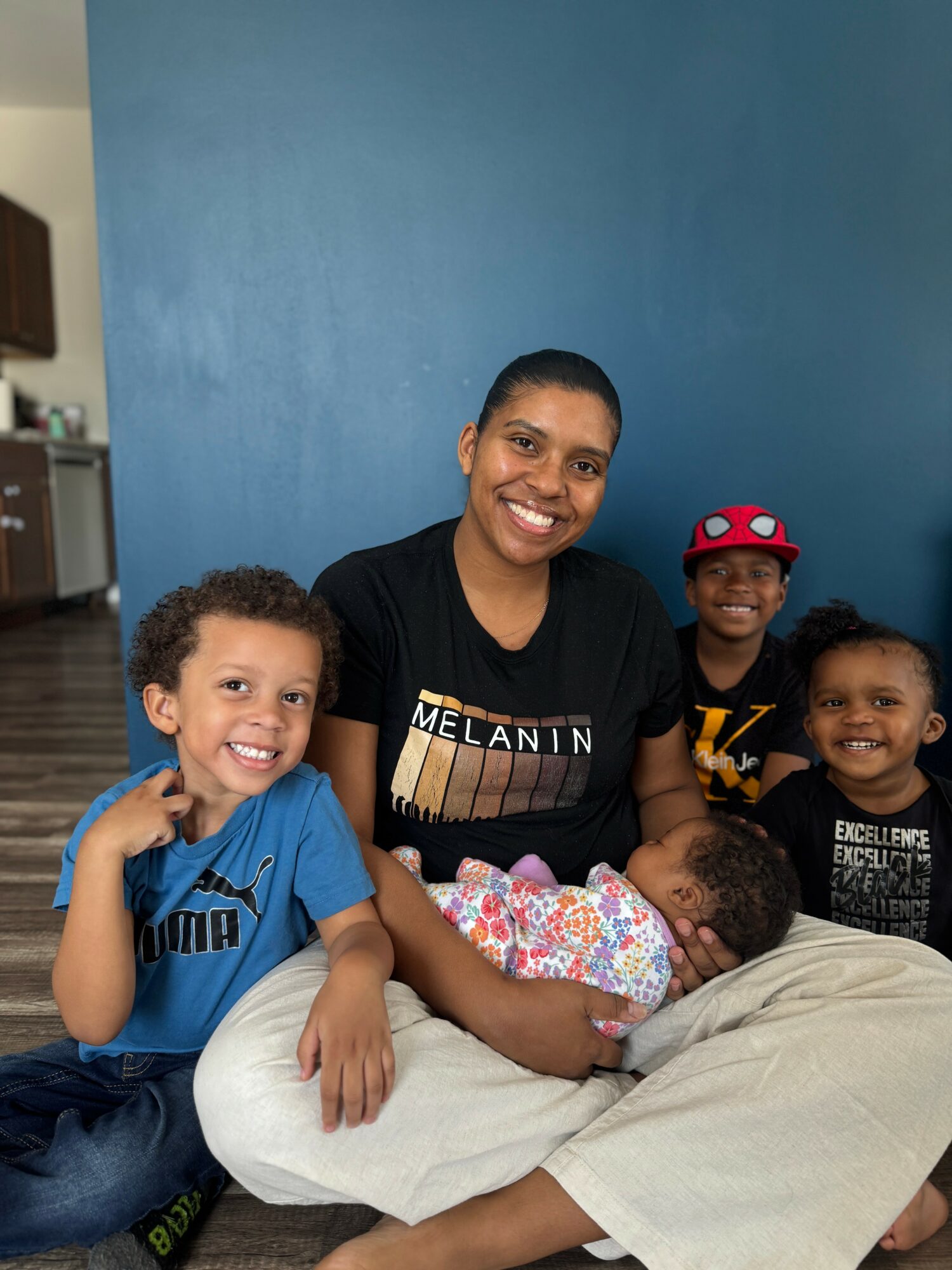 Group of five people, including a woman and four children, sitting together indoors against a blue wall, smiling.