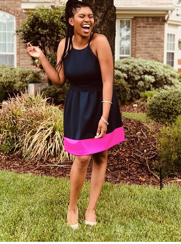 Woman laughing outdoors in a black and pink dress, standing on grass with bushes and a house in background.