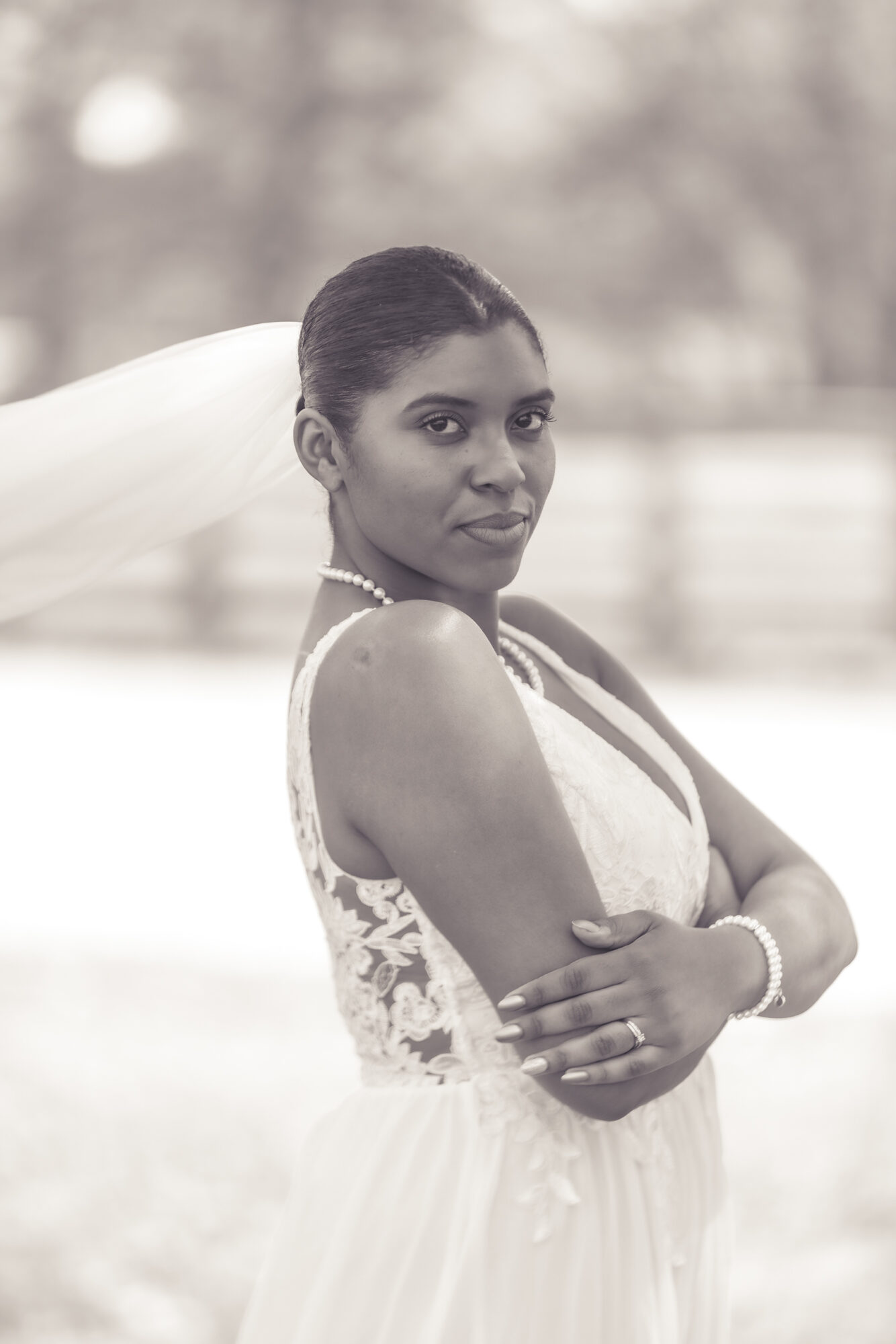 Woman in wedding dress with arms crossed, outdoors, with a veil and pearl necklace, looking at the camera.