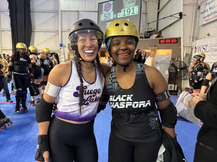 Two smiling women wearing helmets and roller derby gear, standing on a blue rink with other players in the background.
