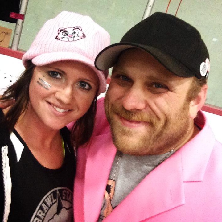 Two people smiling, woman wearing a pink hat with a cat face, man in a black cap and pink jacket, indoors.