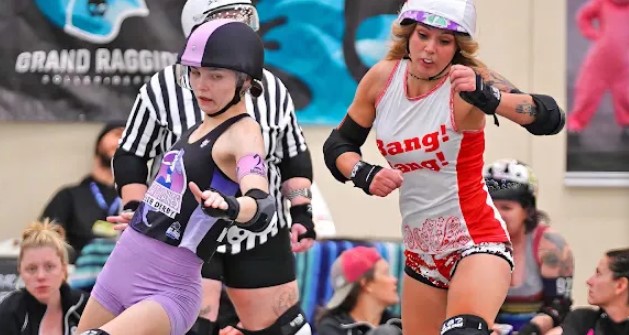 Two young women in roller derby gear, wearing helmets and protective pads, standing in a skating rink with spectators in the background.