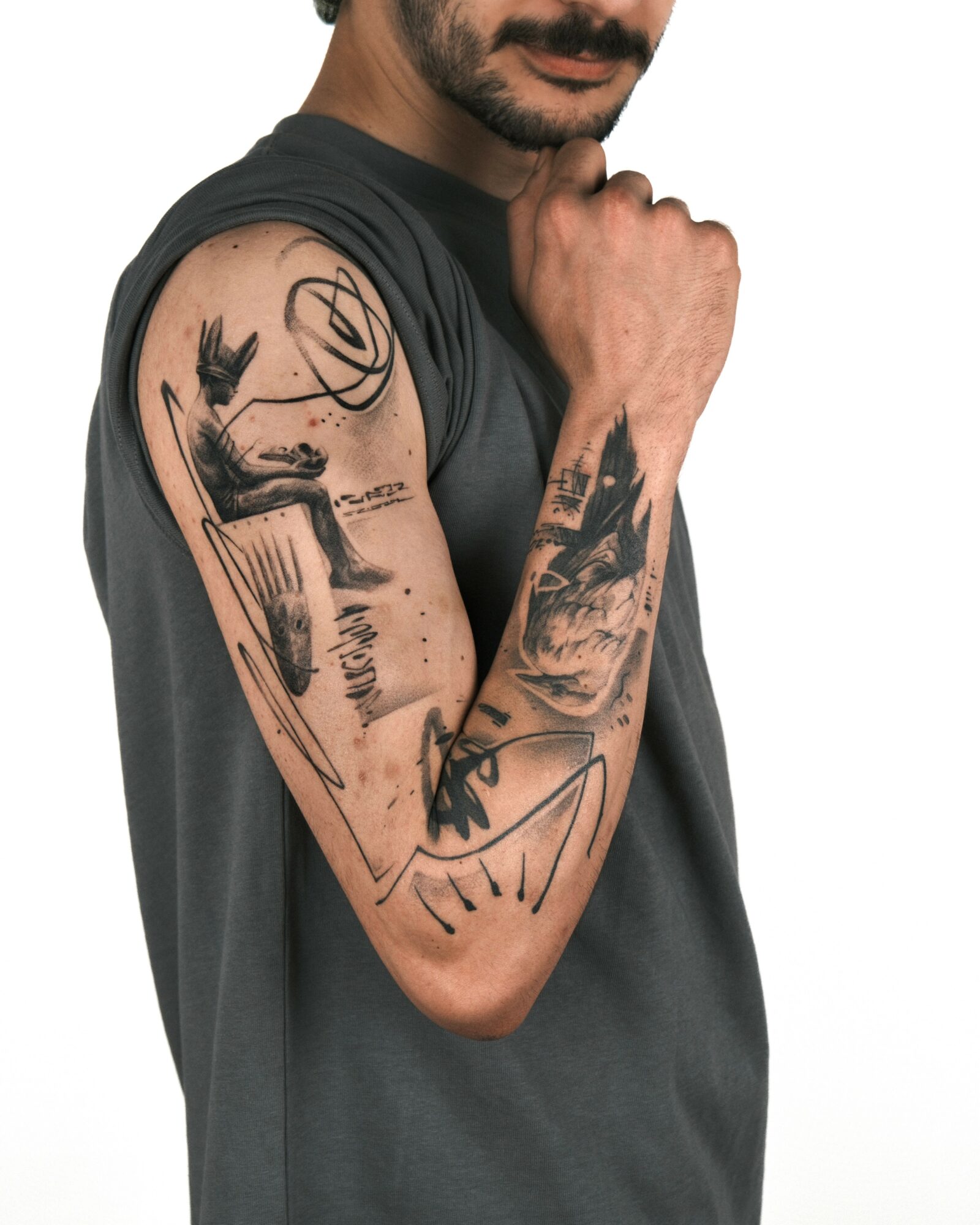 Man with tattoos on his arm, wearing a dark sleeveless shirt, flexing his arm with a fist near his face.