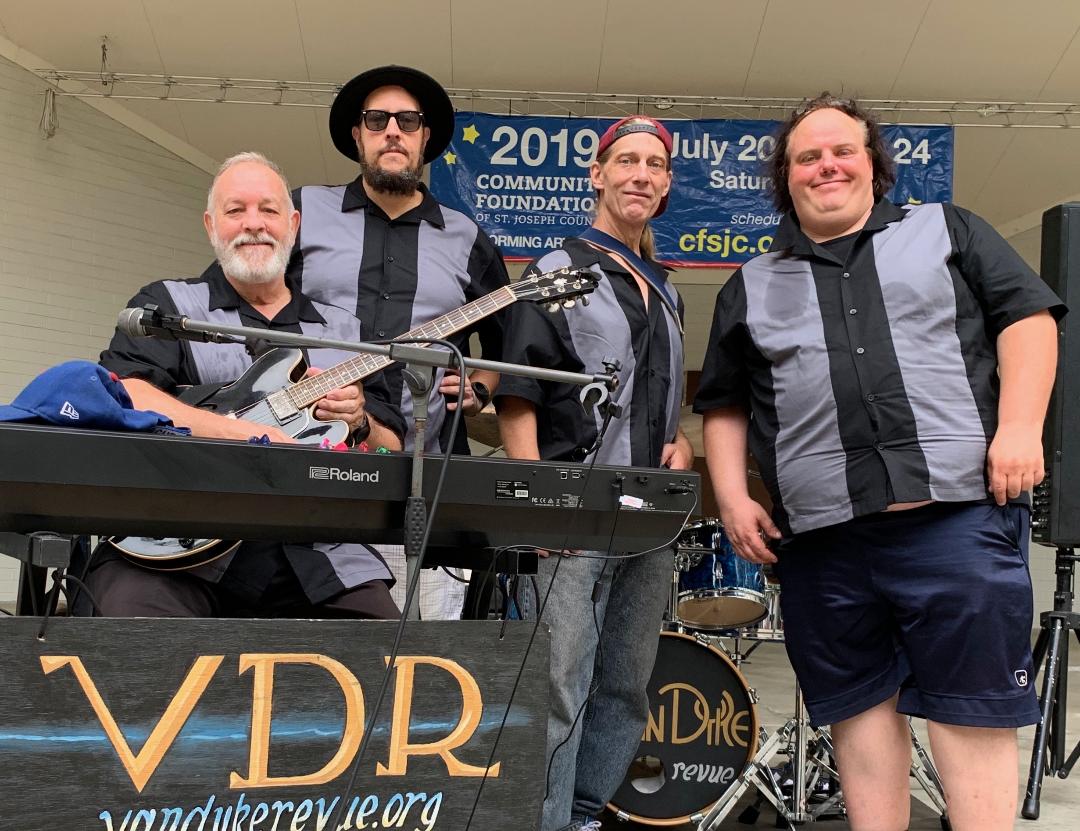 Four people standing outdoors, one playing a guitar, with a banner in the background, and a keyboard in front.