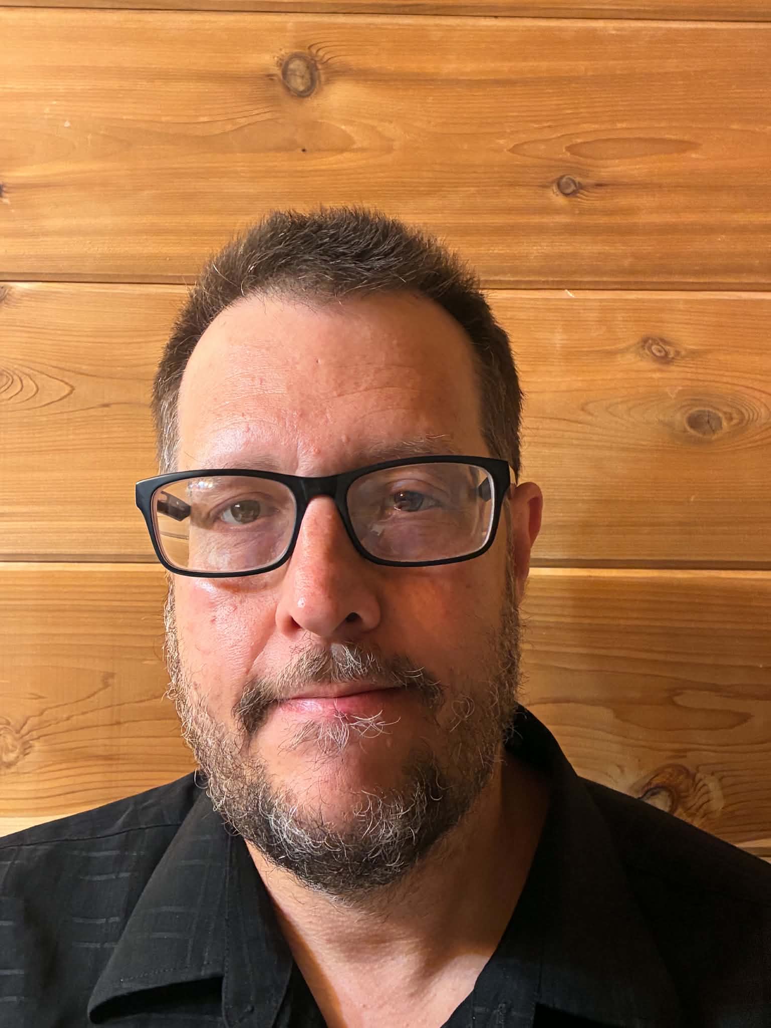 Man with glasses and beard, facing camera, wooden wall background.