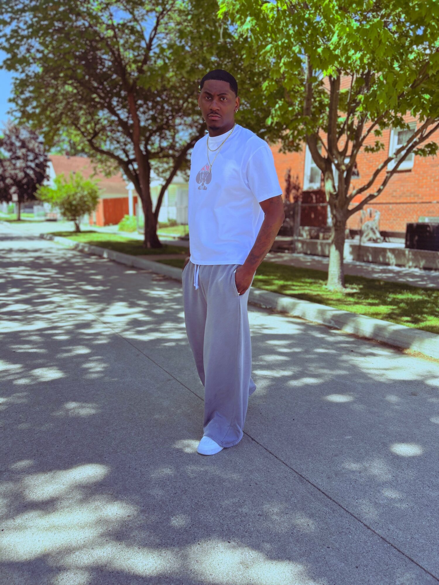 Young man standing outdoors on a sidewalk with trees and buildings in background, wearing a white t-shirt and light-colored pants.