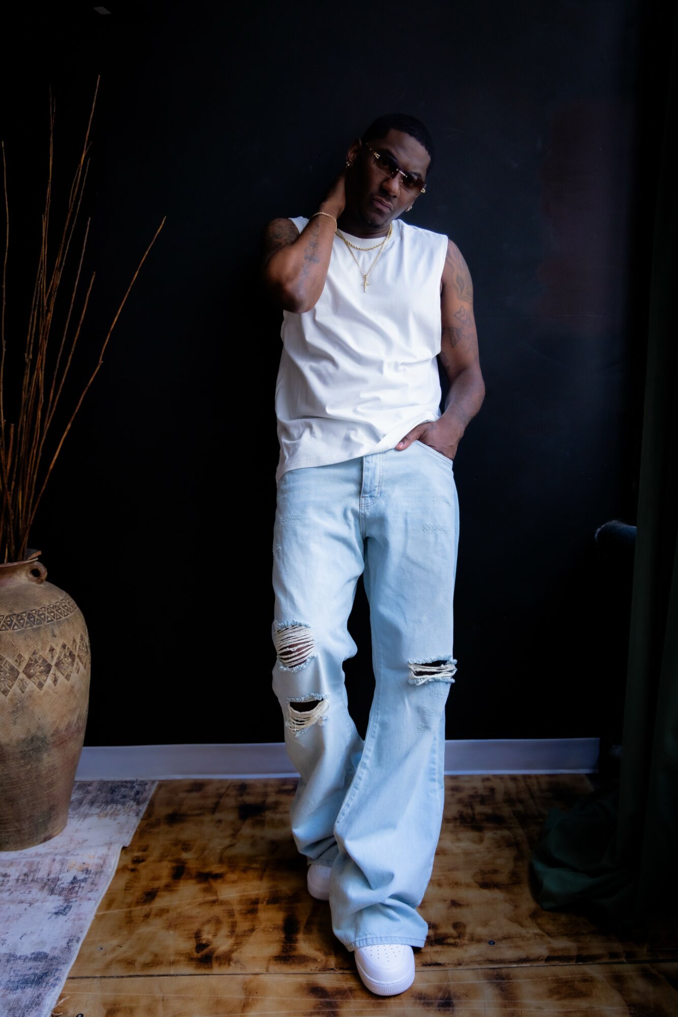 Person standing indoors, wearing a white sleeveless top and ripped jeans, with one hand on neck and the other in pocket.