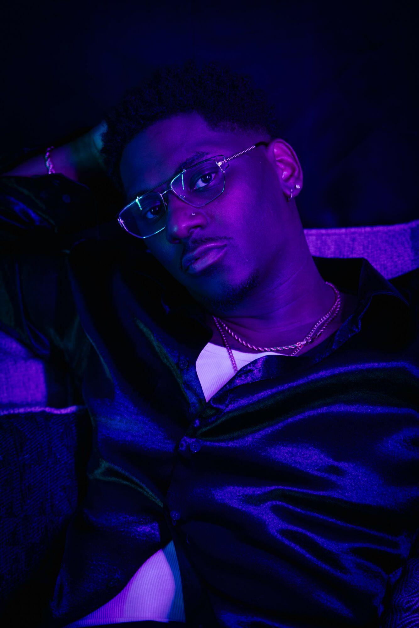 Person wearing glasses and a dark shiny shirt, illuminated by purple lighting, looking at the camera with a relaxed pose.