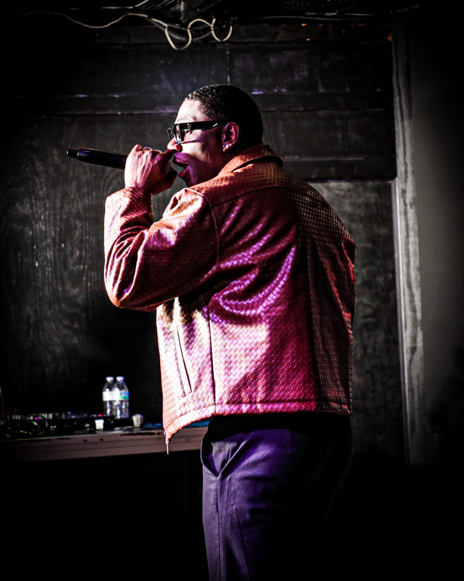 Person wearing sunglasses and a shiny jacket, holding a microphone, standing on stage with a dark background.