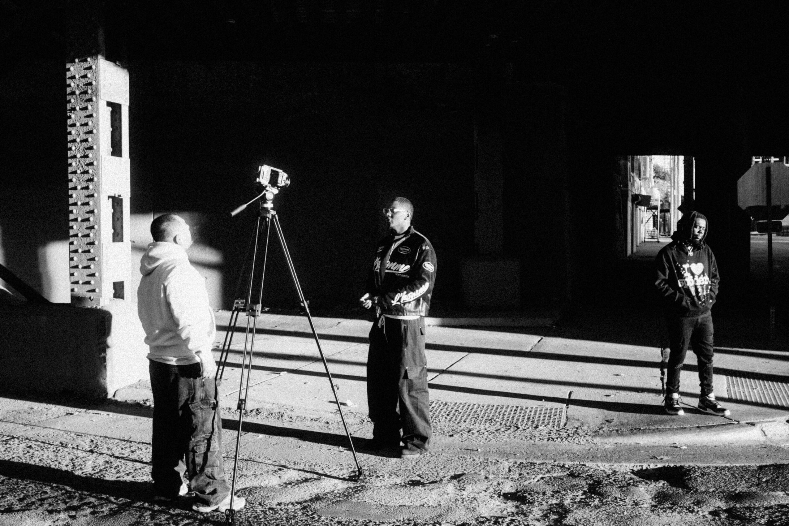 Three people are outdoors near a building, with one person operating a camera on a tripod, and two others standing nearby.