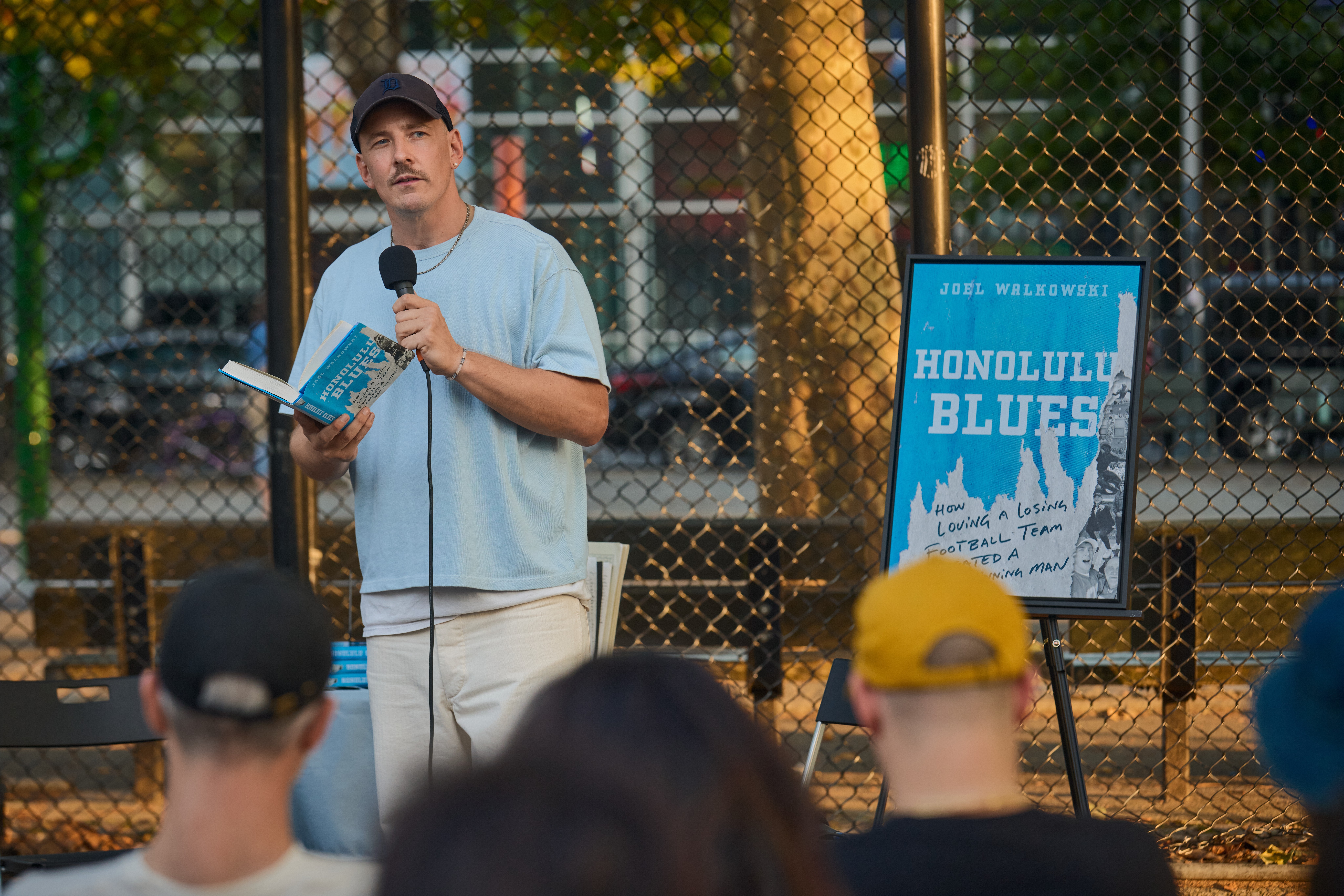 Man speaking into microphone while holding a book outdoors, with a sign reading 'Honolulu Blues' and a group of people listening.
