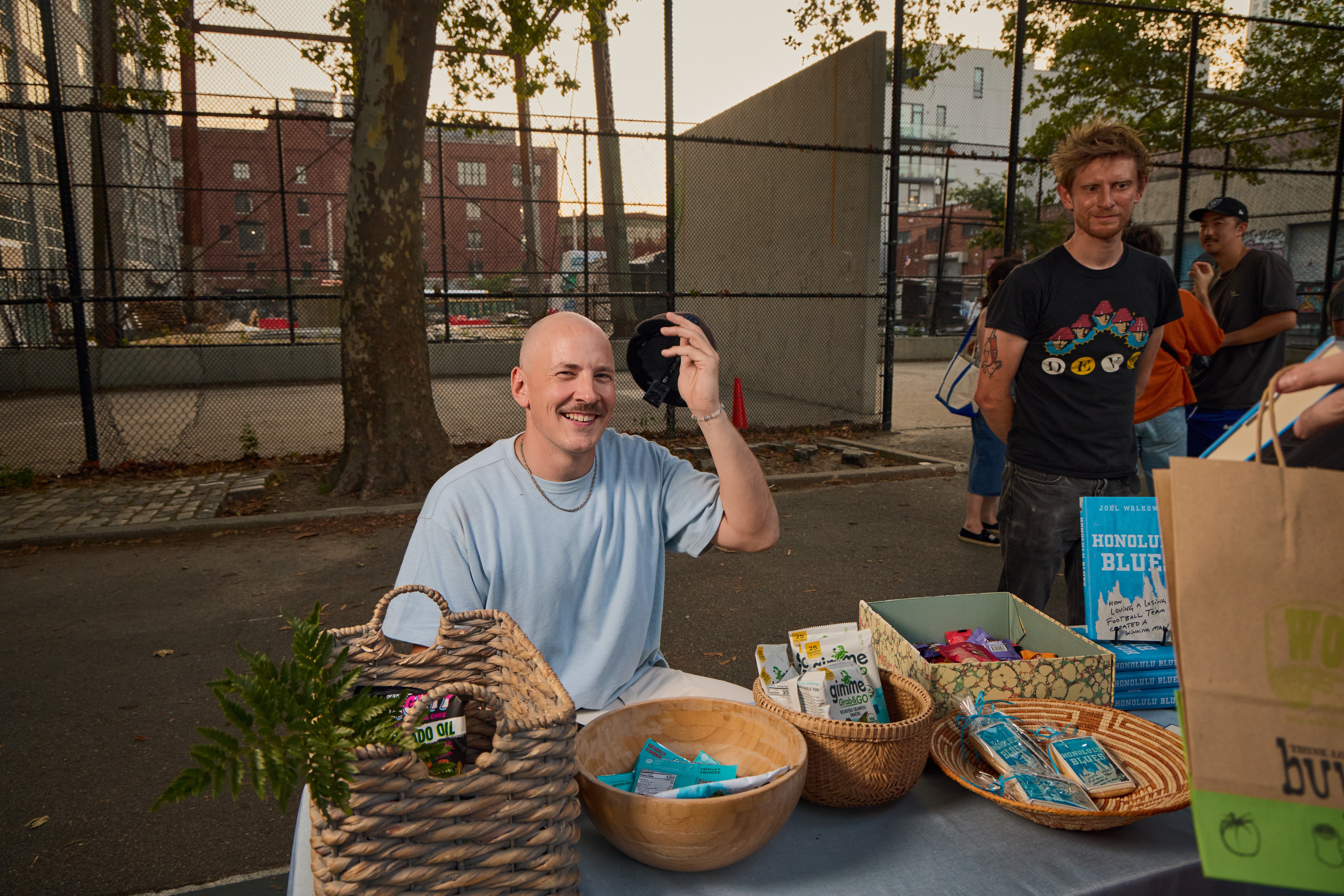 Man smiling and waving at an outdoor event with baskets and a plant on the table, people and trees in the background.
