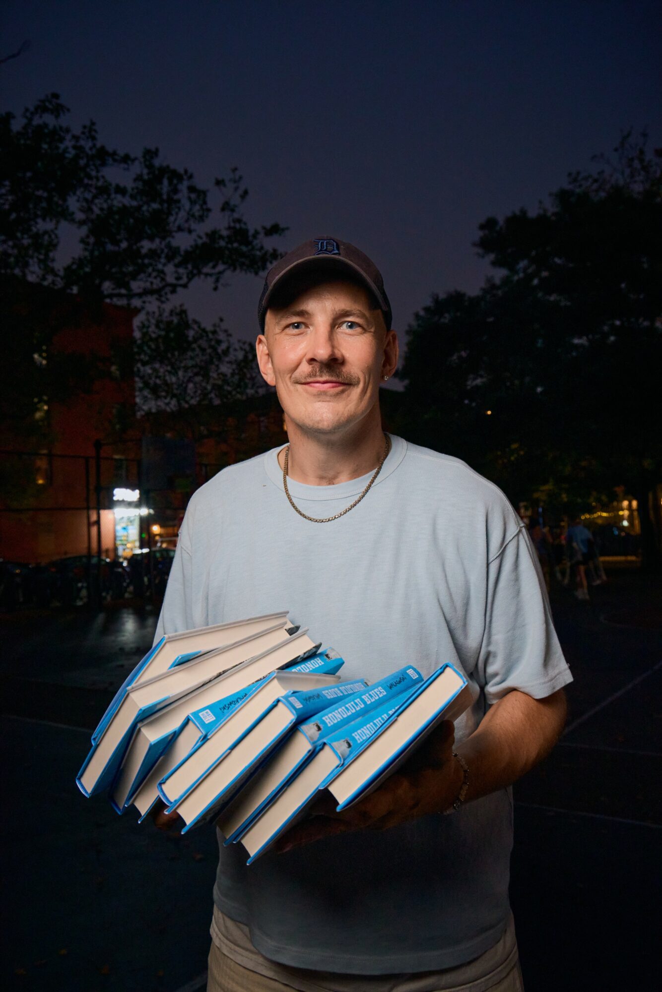 Man holding a stack of books outdoors at night, smiling, wearing a cap and light-colored T-shirt.