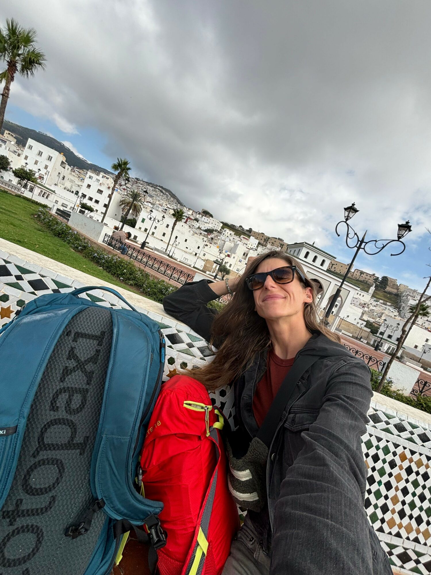 Woman with sunglasses and dark hair resting on a patterned railing with cityscape and cloudy sky behind her.