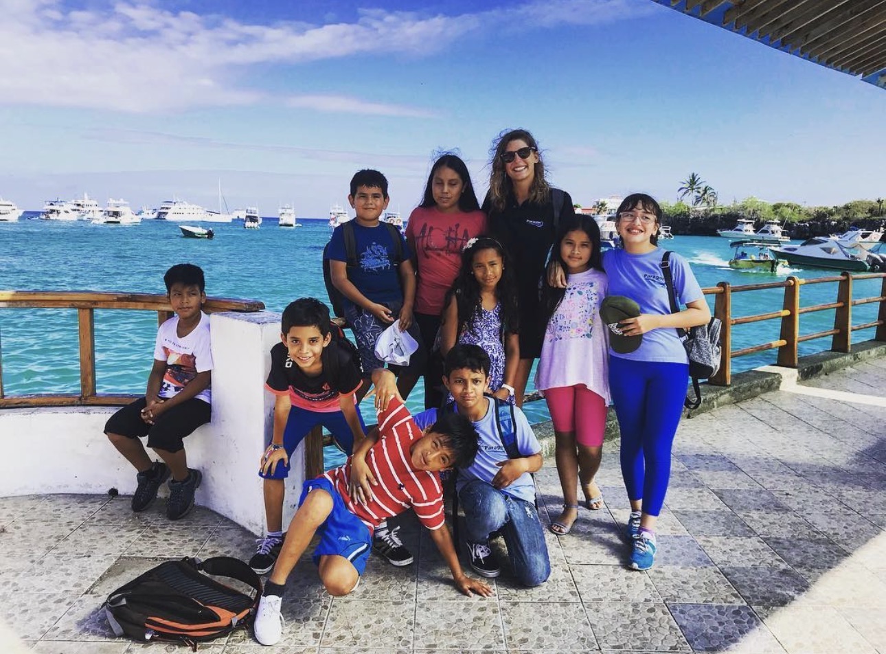 Group of ten people, including children and an adult, posing outdoors near a body of water with boats, under a shaded structure.