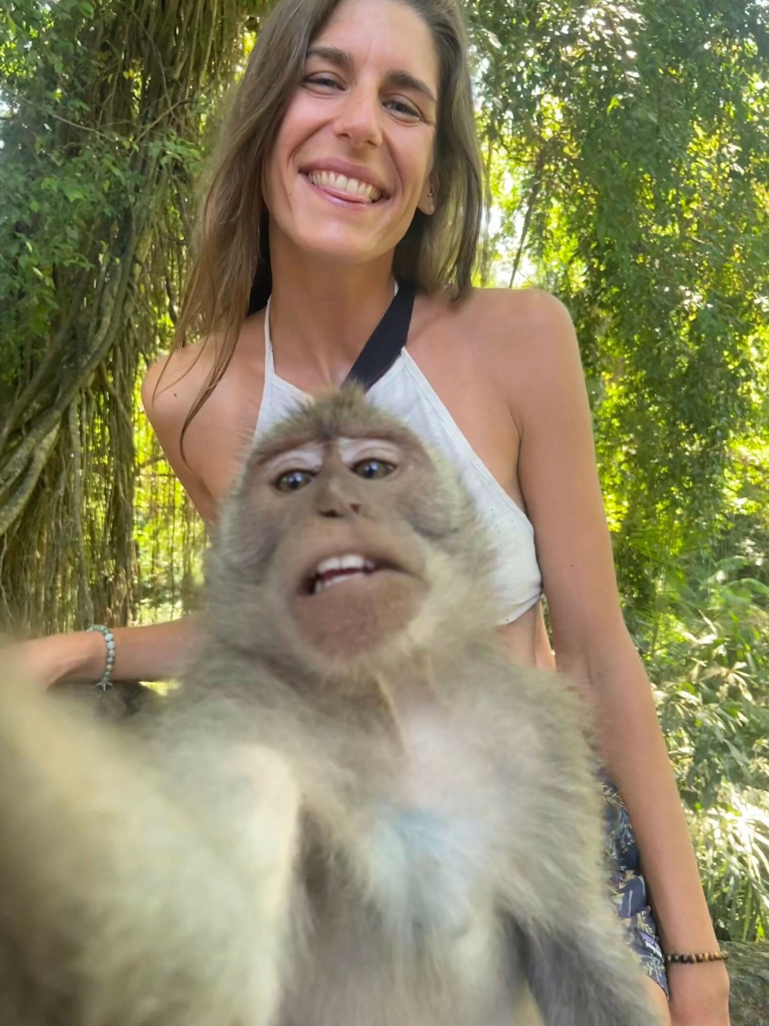 Young woman smiling outdoors with a monkey in front of her, surrounded by green trees.