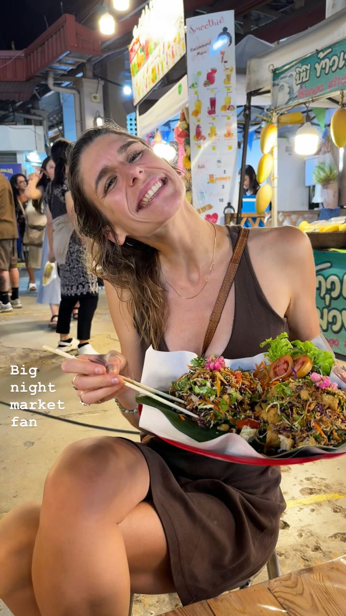 Woman smiling and holding a plate of food at a night market, with colorful signs and people in the background.