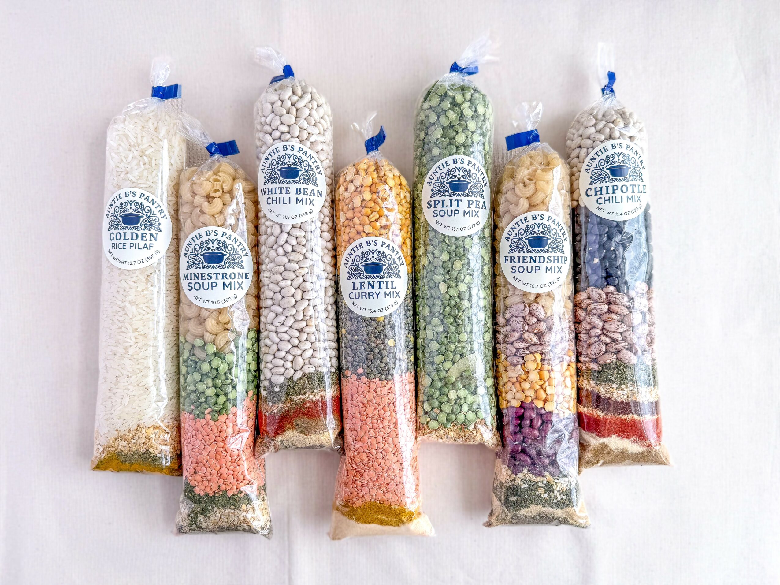 Seven clear plastic bags filled with various dried beans and grains, each with a blue tie and label, arranged in a row.