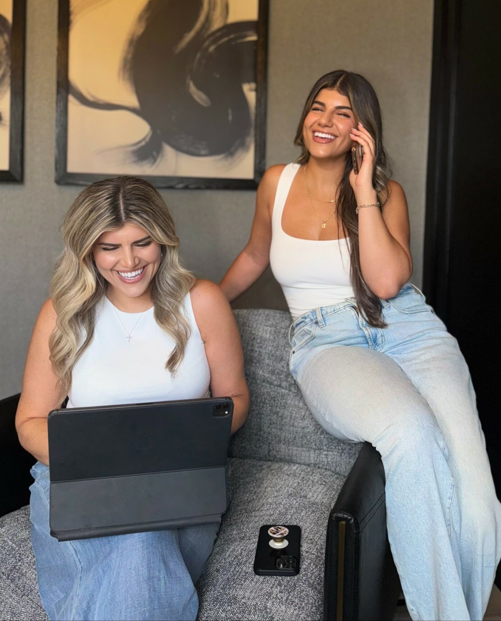 Two women sitting on a gray couch, one using a tablet and the other talking on a phone, smiling.